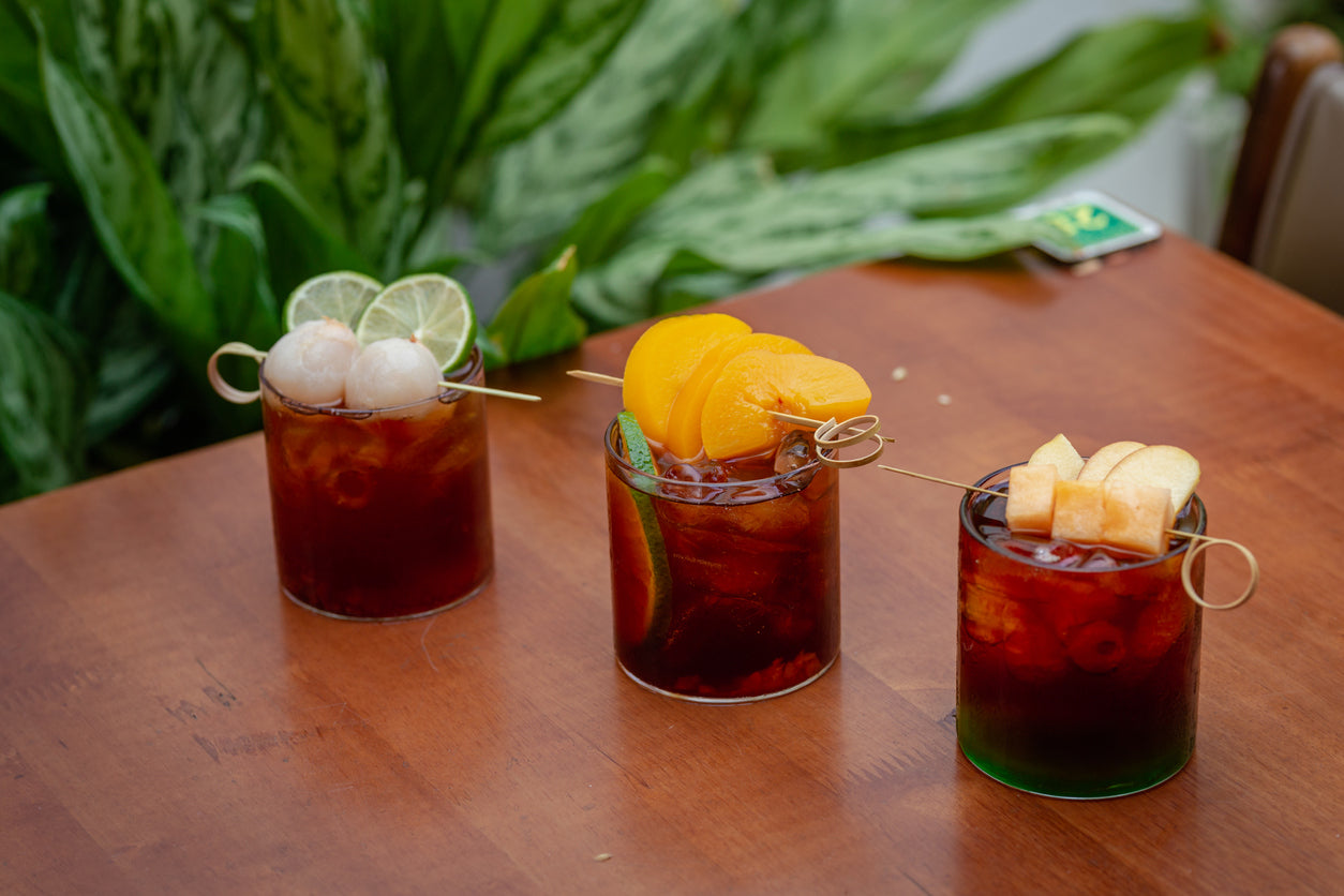 Three cocktails with fruit and ice on a wooden table with greenery in the background