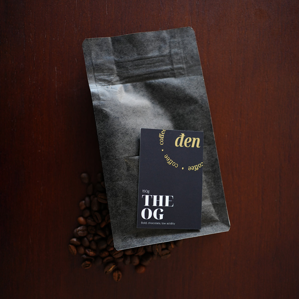 Coffee bag labeled 'den' with a card labeled 'THE OG' on a wooden surface.